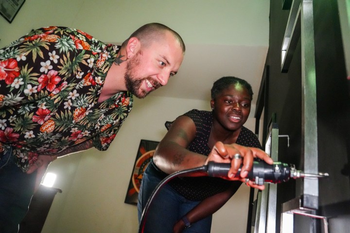 Two people use a power tool in a room, one holding and one assisting.