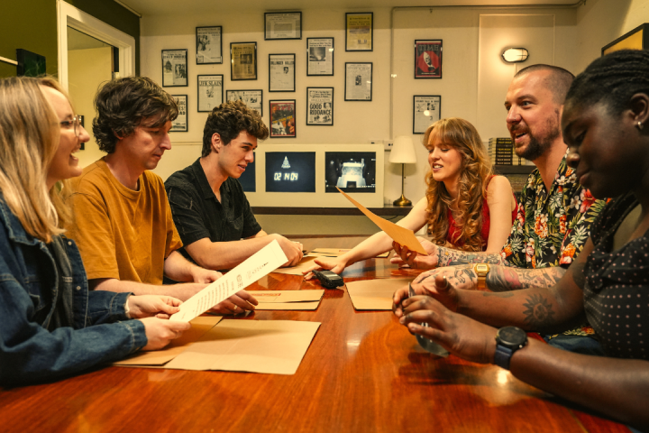 Group of six people at a table discussing and holding papers in a room with framed pictures on walls.